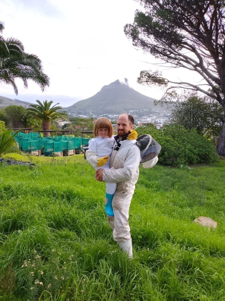 Beekeeper in protective suit holding his daughter in a matching suit in a green field with Lion's Head mountain in the background